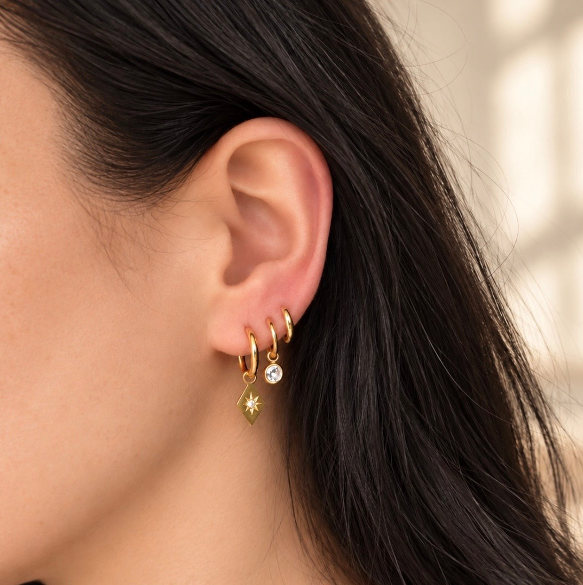 A close-up of a model's ear wearing a stack of gold hoop earrings, including a small gold star charm and a clear crystal accent, against a softly blurred light background.