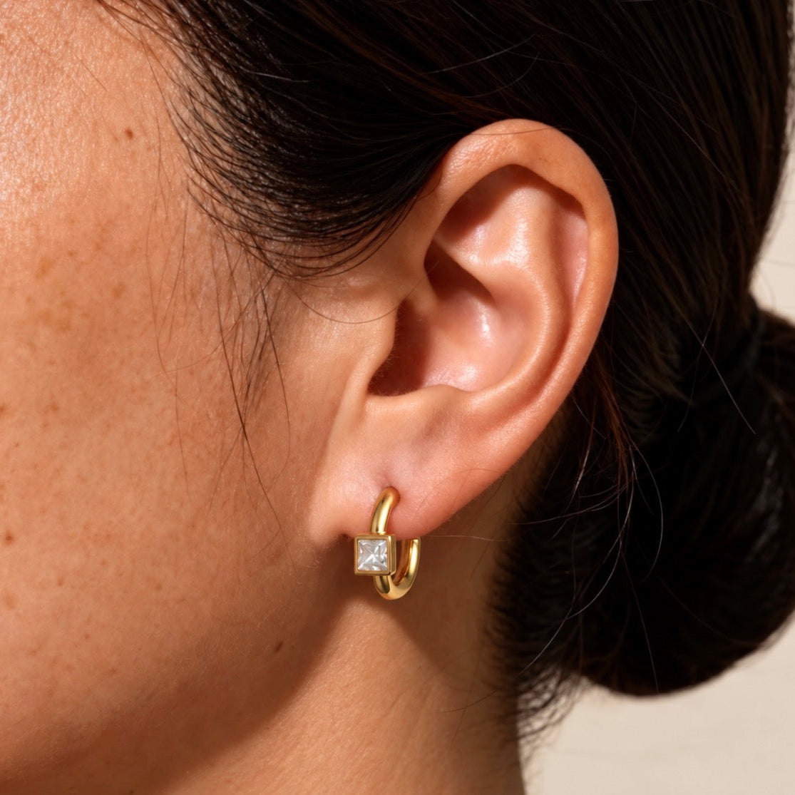 A close-up of a person's ear wearing a chunky gold huggie hoop earring with a square clear gemstone at the hinge, against a light neutral background.