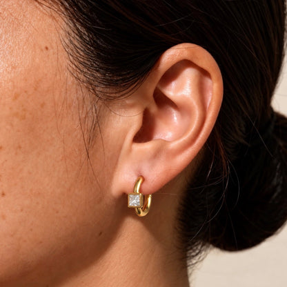 A close-up of a person's ear wearing a chunky gold huggie hoop earring with a square clear gemstone at the hinge, against a light neutral background.