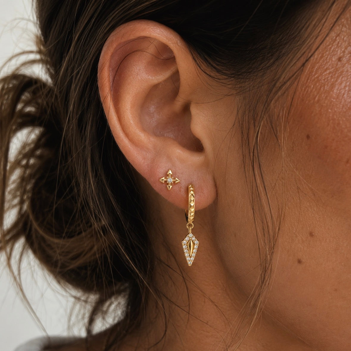 A close-up of a model's ear wearing a pair of gold earrings, including a small cross-shaped stud earring and a huggie hoop with a dangling geometric charm.