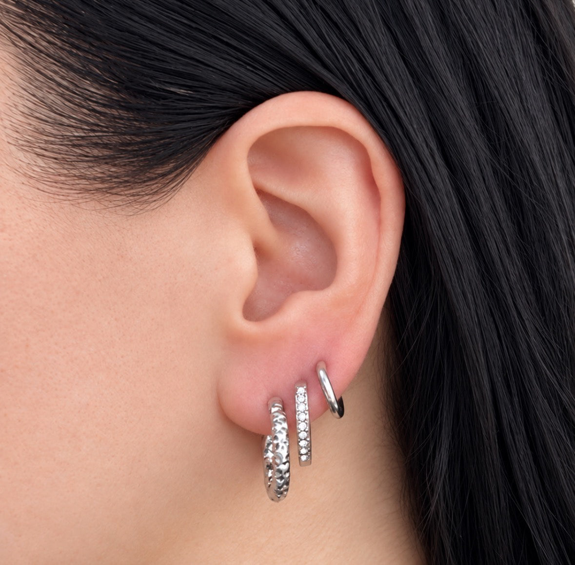 A close-up of a person's ear wearing three silver huggie hoop earrings, including one textured hoop and two small pavé-set hoops, against dark hair and a neutral background.