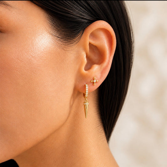 A close-up of a model's ear wearing a delicate gold hoop earring with a small diamond-studded spike dangle and a matching small gold stud.
