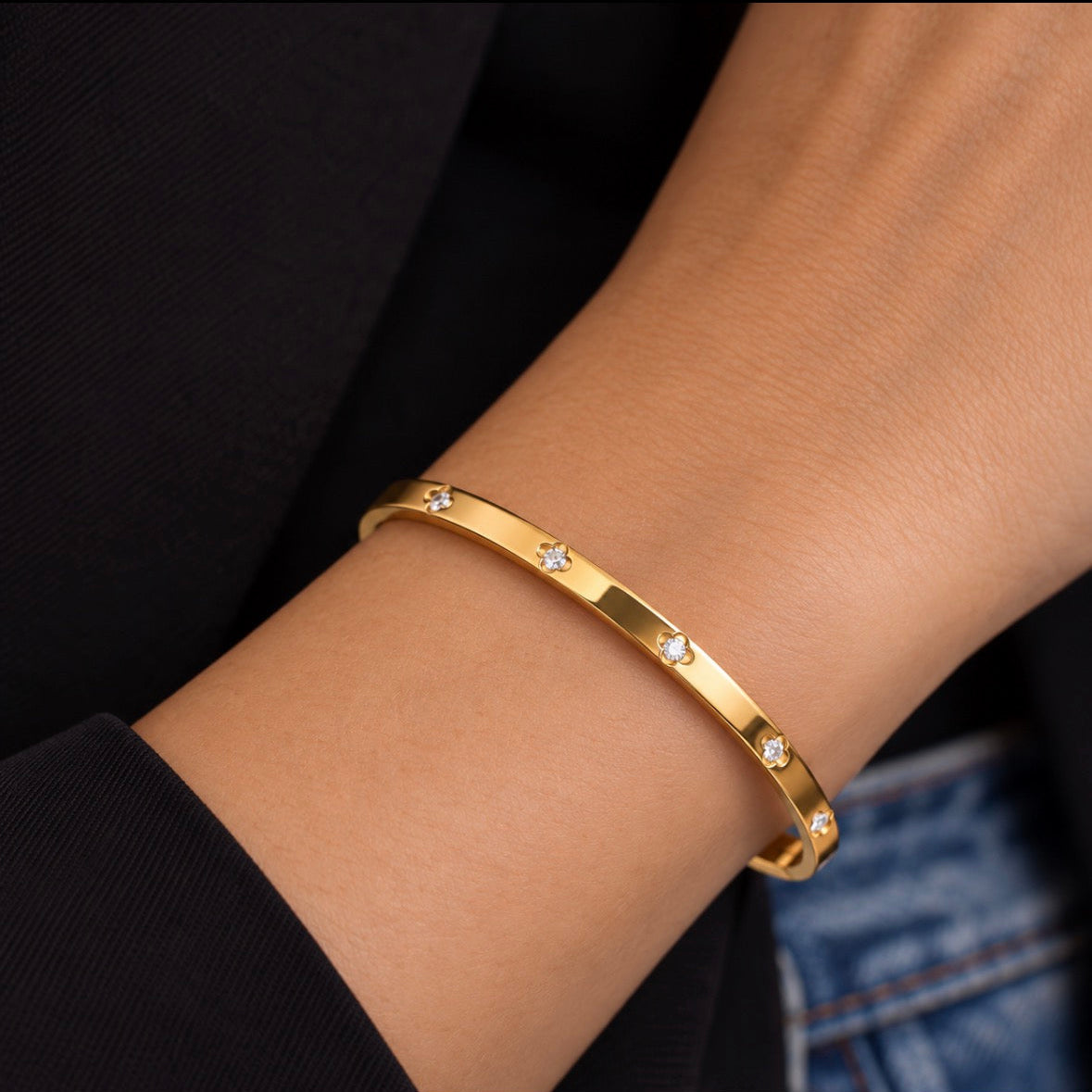 A gold bangle bracelet with small star-shaped clear stone accents, worn on a person's wrist against a dark black top and blue denim background.