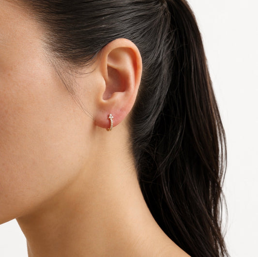 A close-up of a model's ear wearing a small gold huggie hoop earring adorned with tiny clear stones, set against a clean white background.