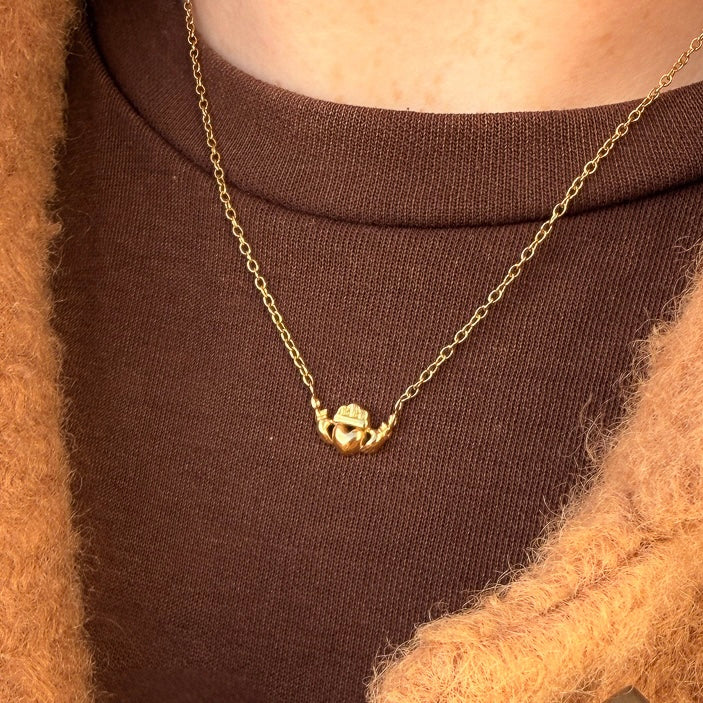 Gold necklace with a claddagh pendant worn by a person in a brown sweater.