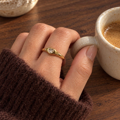 A close-up lifestyle shot of a hand wearing a delicate gold ring with a small heart-shaped clear gemstone, resting near a ceramic coffee cup on a wooden table.