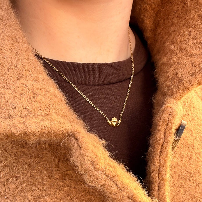 Close-up of a person wearing a gold claddagh necklace. partially visible under a brown coat.