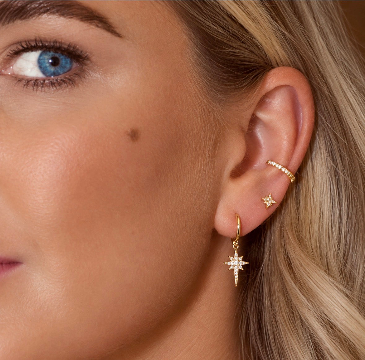 Close-up of a woman's ear wearing gold jewellery, including a dangling starburst earring, a small stud, a cz ear cuff at conch ear piercing placement 
