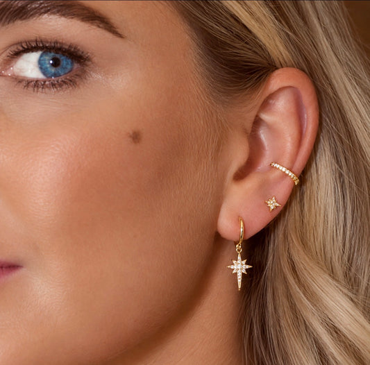 Close-up of a woman's ear wearing gold jewellery, including a dangling starburst earring, a small stud, a cz ear cuff at conch ear piercing placement 