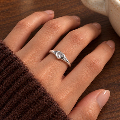 A silver ring with a heart-shaped clear gemstone set on a person's ring finger, shown resting on a wooden table next to a ceramic mug.