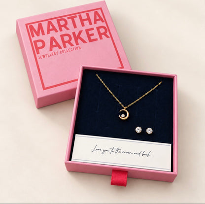 A gold necklace and matching stud earrings presented in an open pink Martha Parker gift box with a dark blue velvet insert, alongside the pink branded box lid on a light beige background.