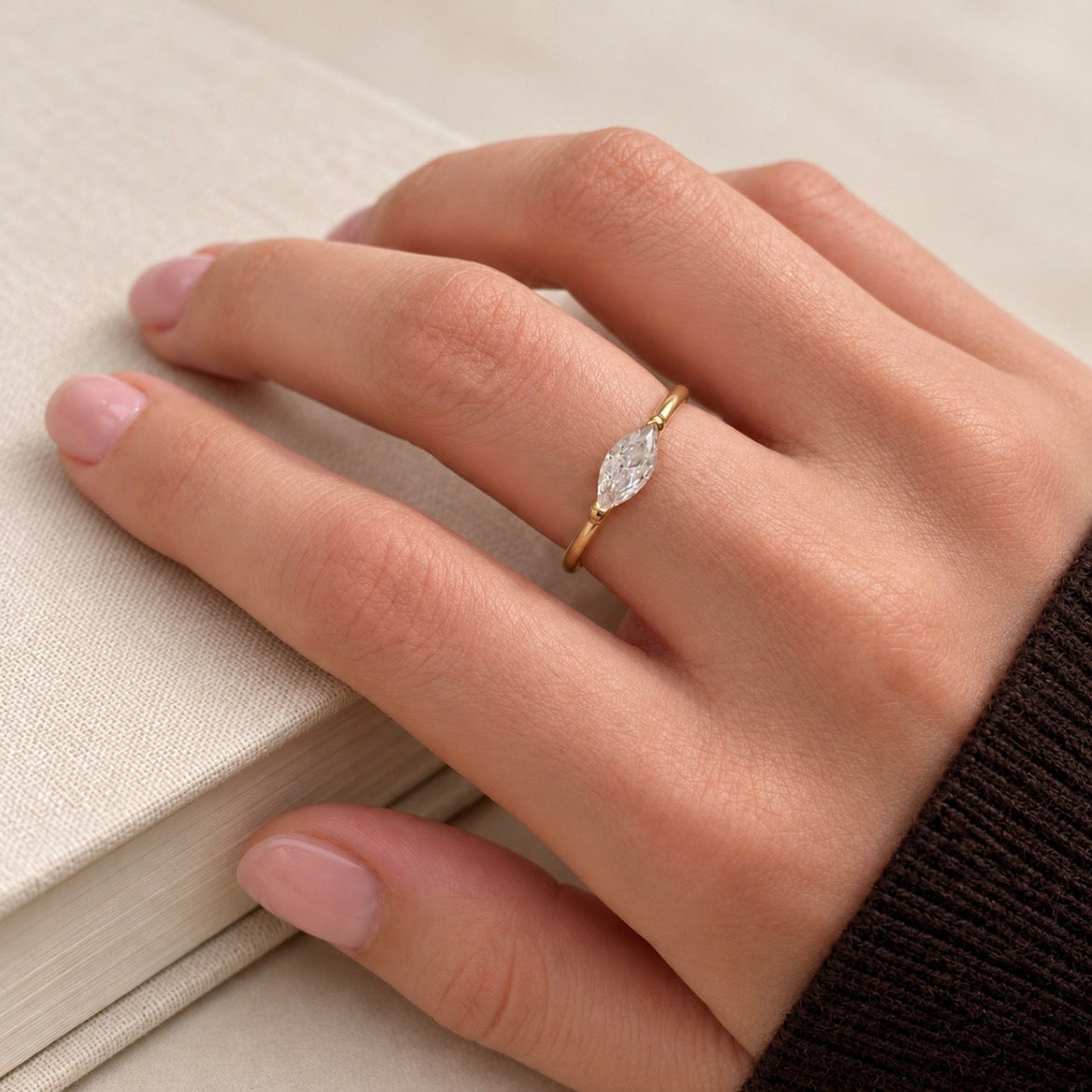 A close-up of a hand wearing a delicate gold ring with a marquise-cut clear gemstone, resting on a textured beige book.