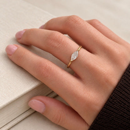 A close-up of a hand wearing a delicate gold ring with a marquise-cut clear gemstone, resting on a textured beige book.