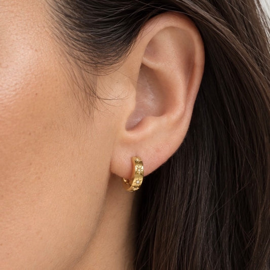 A close-up of a model's ear wearing a small gold huggie hoop earring with textured detailing.