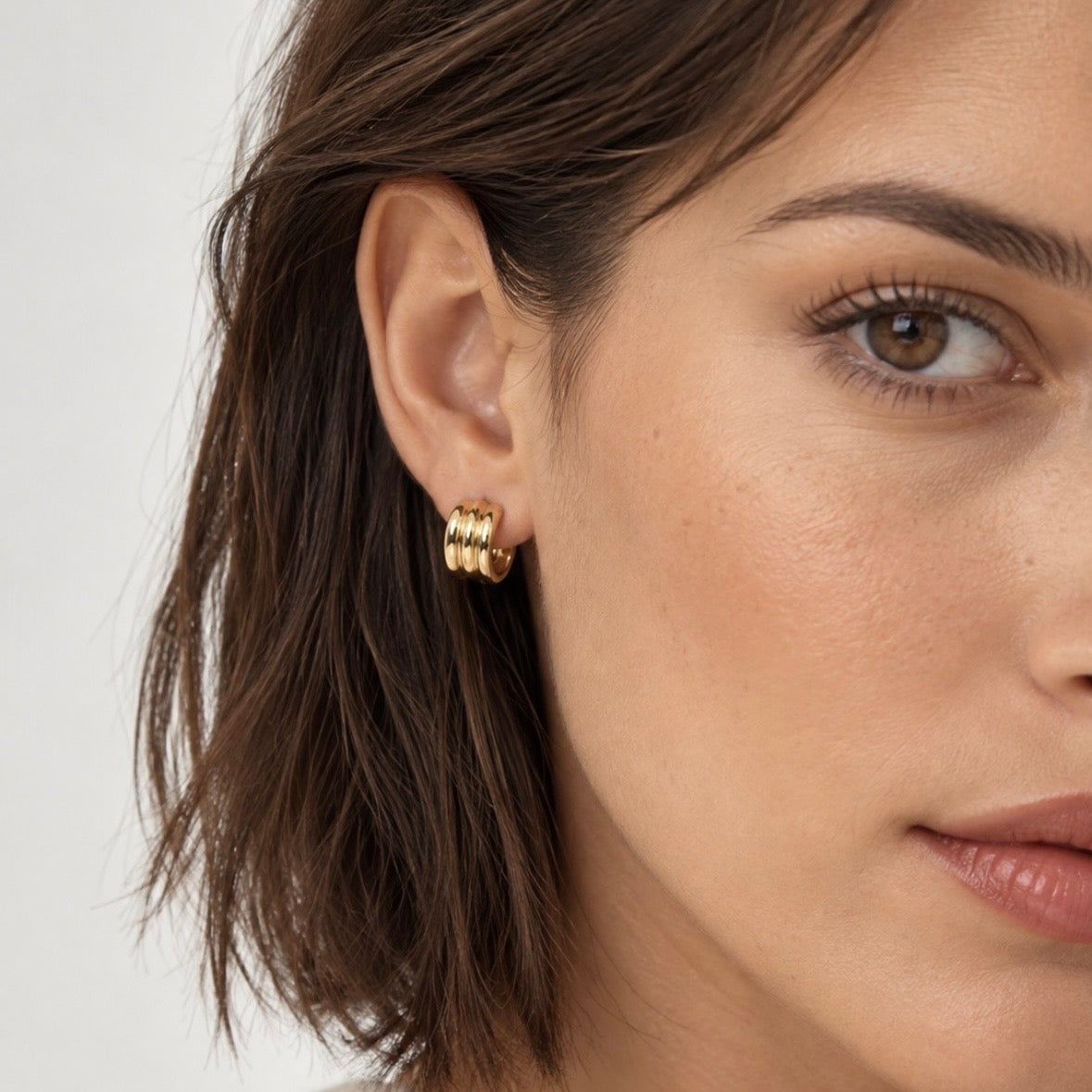 A close-up of a woman's ear wearing a small gold hoop earring with three stacked textured hoops, set against a clean light gray background.