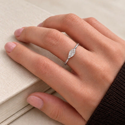 A close-up of a hand wearing a delicate silver ring with a marquise-cut clear gemstone, resting on a textured beige book.