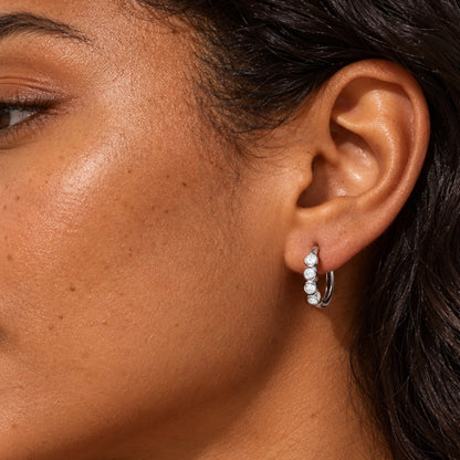 model wearing a silver hoop earrings adorned with four round-cut clear stones in bezel settings.