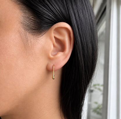 A close-up of a person's ear wearing a small gold safety pin huggie hoop earring, with dark hair and a softly blurred indoor background.