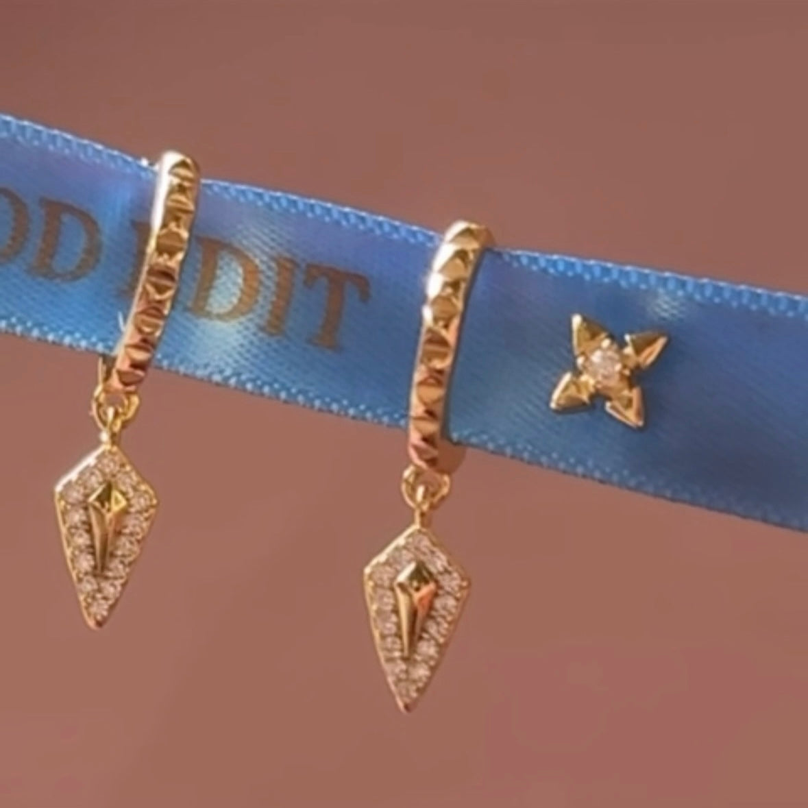 Gold earrings with intricate designs on a blue ribbon with visible text.