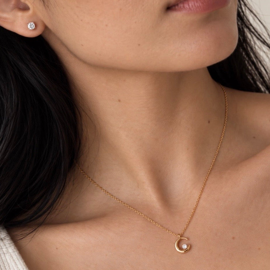 A close-up lifestyle shot of a model wearing a delicate gold crescent moon necklace with a small white cubic zirconia at its center, paired with a matching gold stud earring.