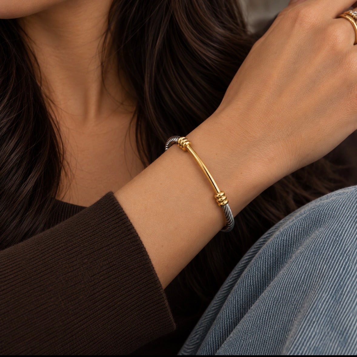 A close-up of a woman's wrist wearing a delicate gold and silver-toned bangle bracelet, with her hand resting near her face and dark hair visible.