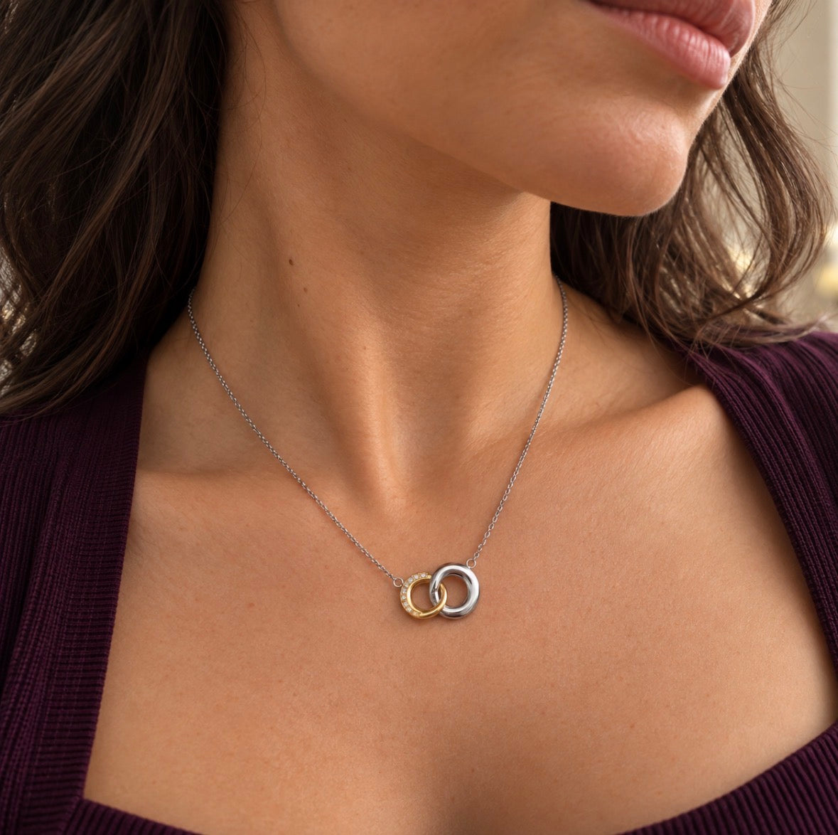 A close-up of a woman's neck and chest wearing a delicate silver chain necklace with two interlocking ring pendants, one silver and one gold, against a dark purple ribbed top.