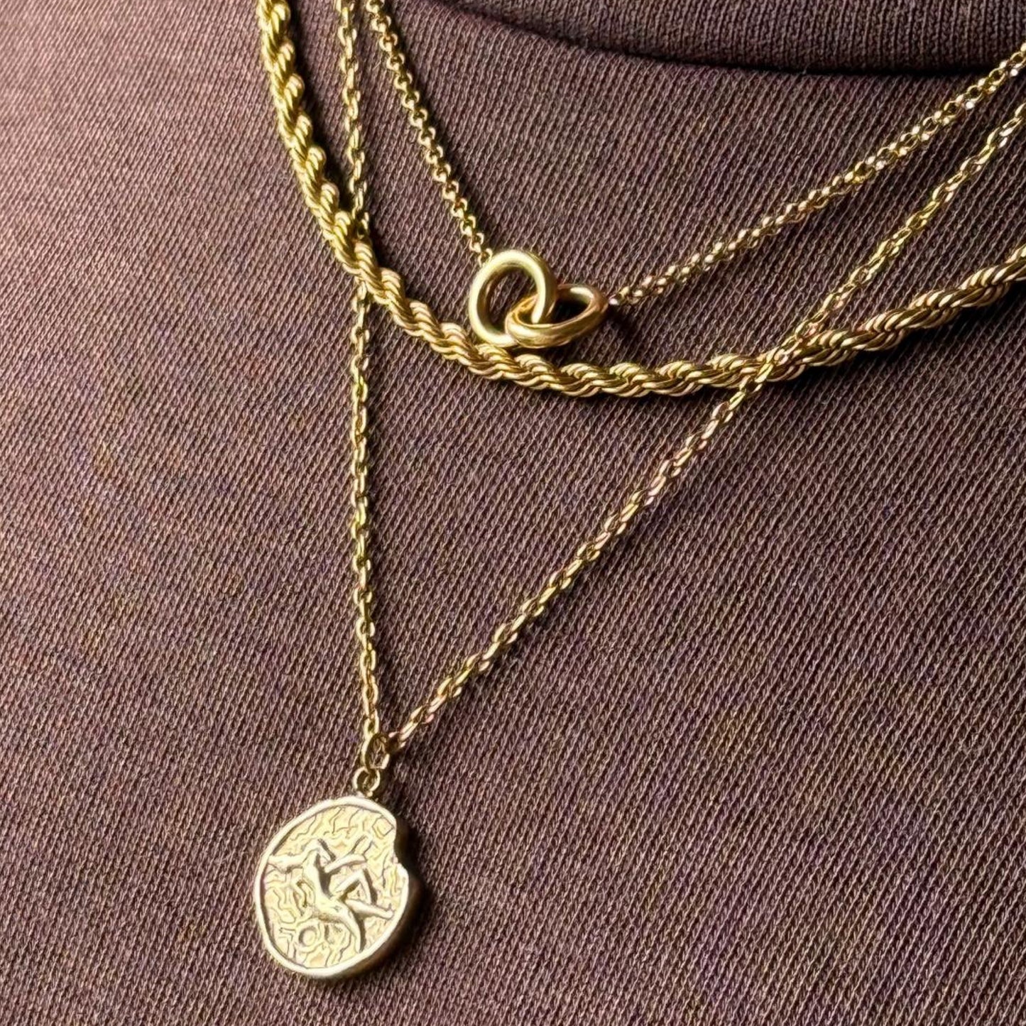 Gold necklaces with a zodiac coin pendant layered with rope chain necklace and double ring necklace on a model brown fabric background