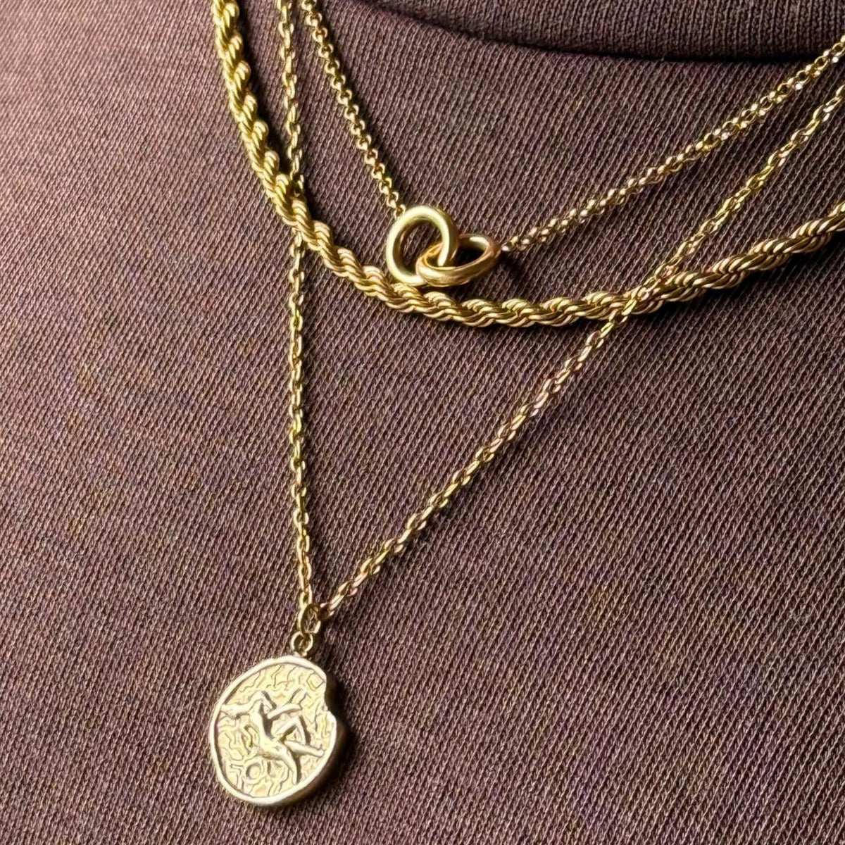 Gold necklaces with a zodiac coin pendant layered with rope chain necklace and double ring necklace on a model brown fabric background
